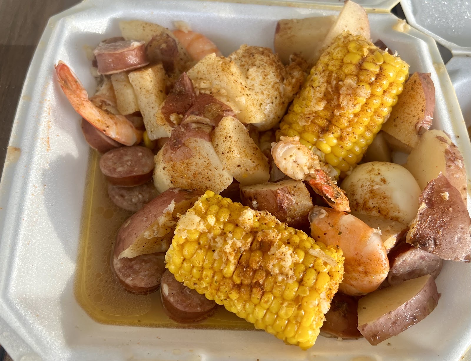 A takeout tray of seafood boil: boiled shrimp, red potatoes, smoked sausage and corn on the cob, dusted with seasoning