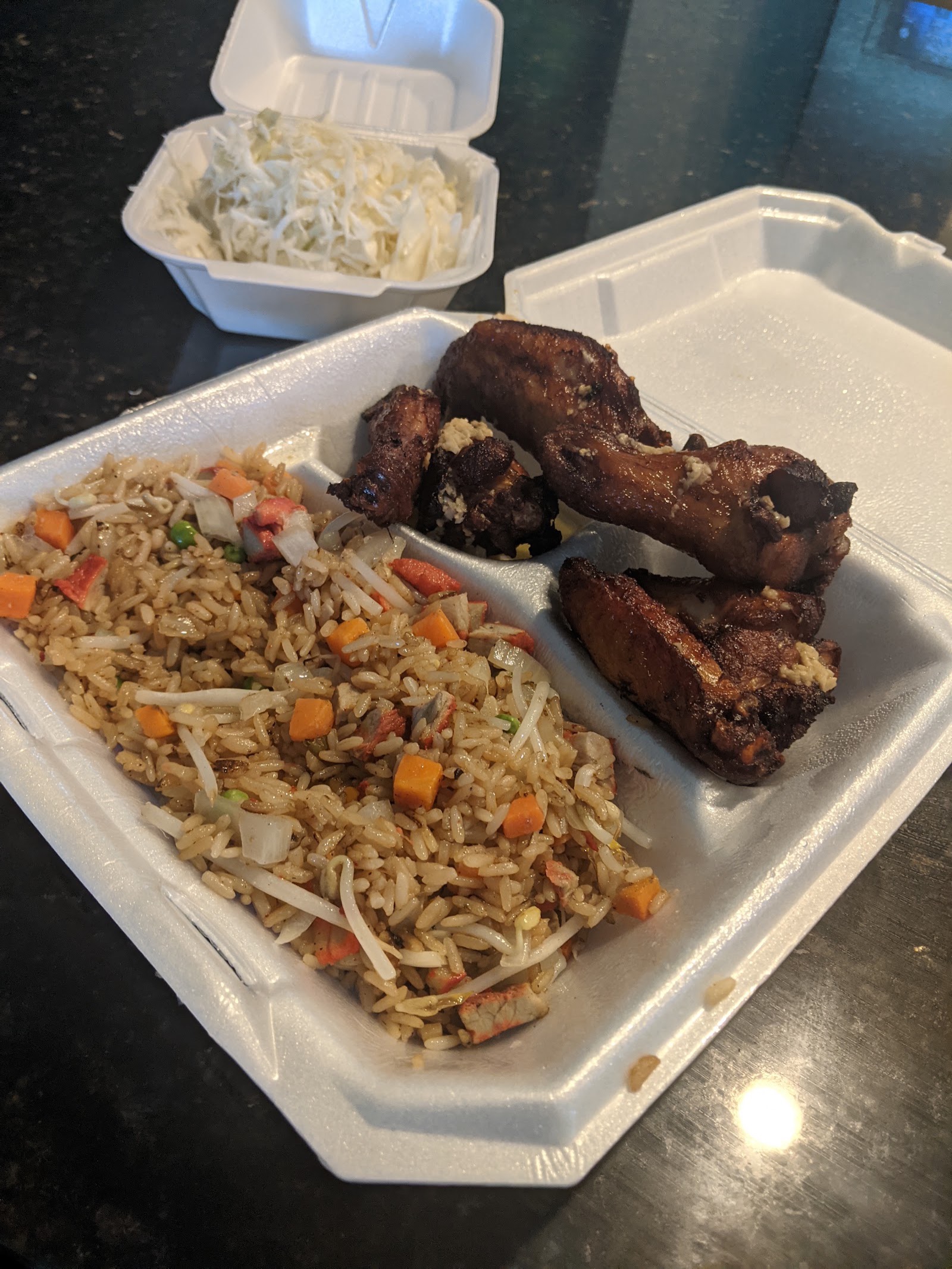 A takeout tray of fried chicken wings beside stir-fried rice with vegetables, with a cup of coleslaw